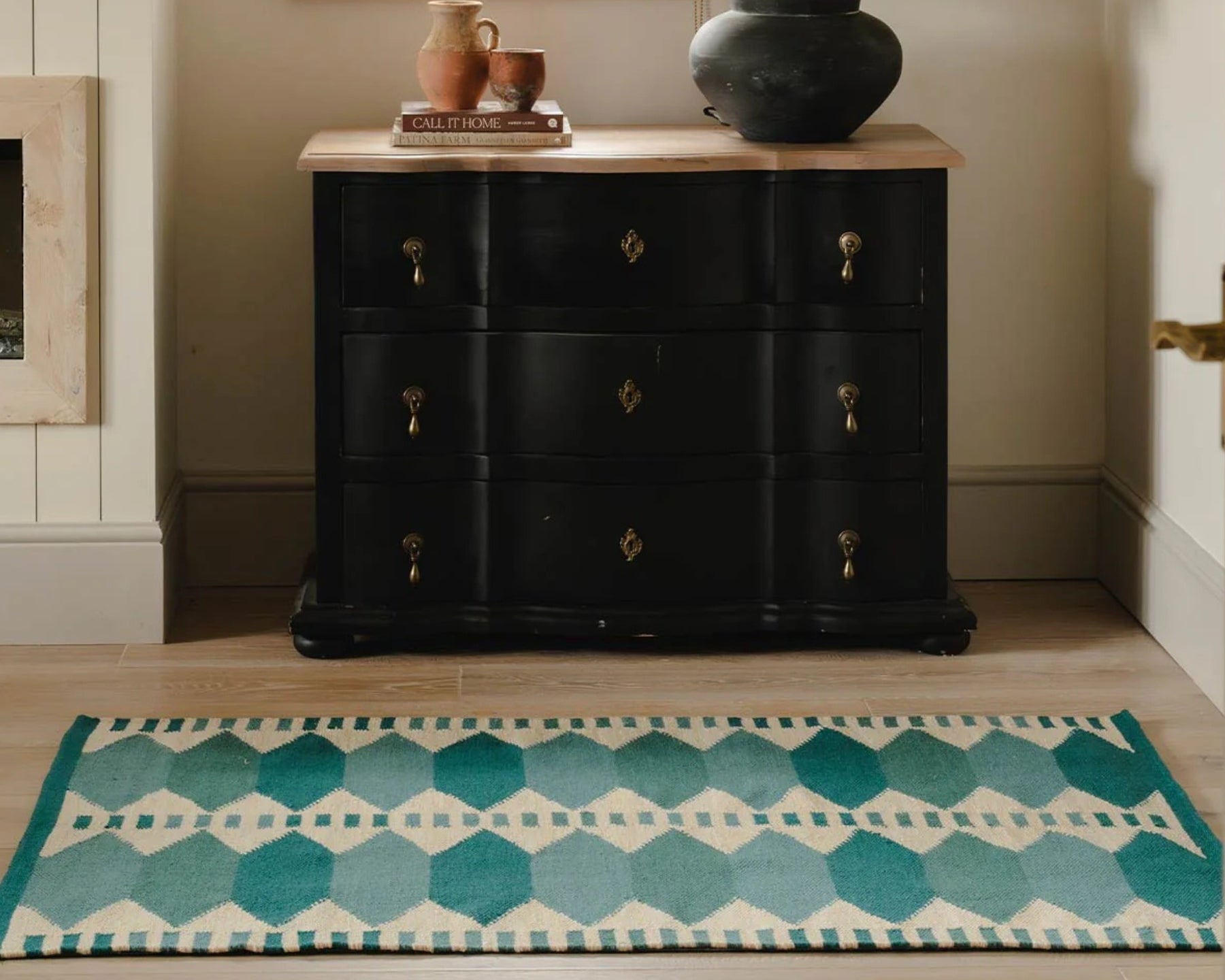 A black wooden chest of drawers with brass handles stands in a styled room corner beside a window, topped with a lamp and decorative objects. A teal and cream patterned rug is visible in the foreground.